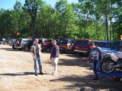 1979 Ford Bronco - Several SUVs are parked in a dirt lot with trees in the background, with people standing around.
