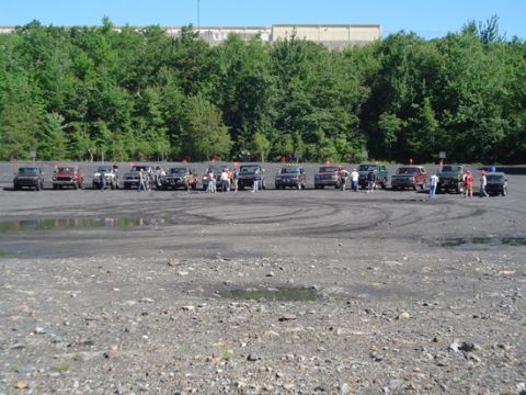 1979 Ford Bronco - A group of off-road vehicles are gathered in an open, gravelly area with a line of trees behind them.