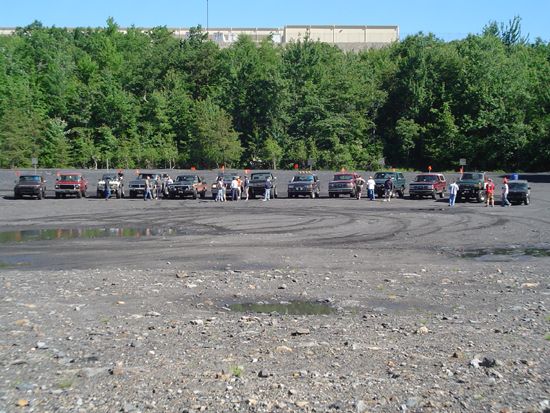 Several SUVs and trucks parked in a dirt lot with trees and a building in the background