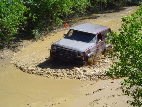 1979 Ford Bronco - A maroon Ford Bronco navigates a muddy off-road trail, splashing water and mud.