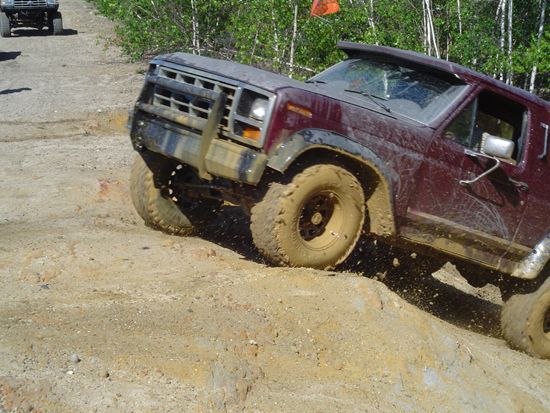 Maroon off-road vehicle with large tires climbing a muddy dirt hill. Another vehicle is visible in the background