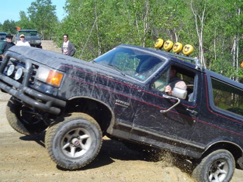1979 Ford Bronco - This black SUV, equipped with smiley face lights, navigates a challenging off-road course, kicking up dirt.