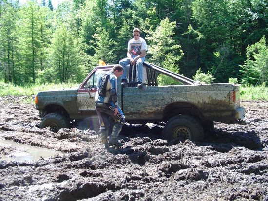 Muddy pickup truck stuck in deep mud with two people standing nearby. One person sits in the truck bed