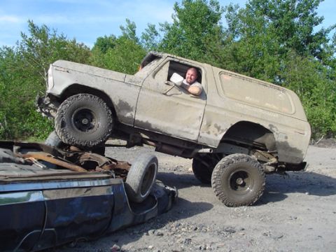 1979 Ford Bronco - A man drives a lifted, mud-splattered Ford Bronco over a flipped-over vehicle in a dirt lot.
