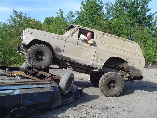 Mud-covered 1978 Ford Bronco climbing over a wrecked car
