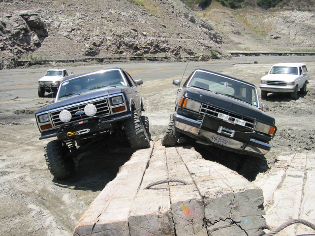 Two dark lifted Ford Broncos climbing over a large log obstacle off-road
