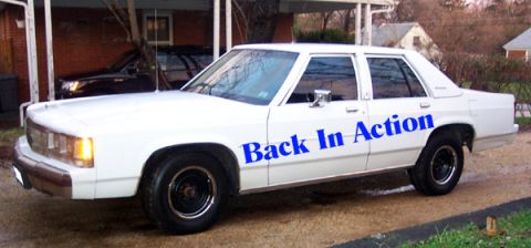 1991 Ford Crown Victoria LTD - A white sedan with blue lettering on its side is parked in a driveway, with another dark vehicle visible behind it.