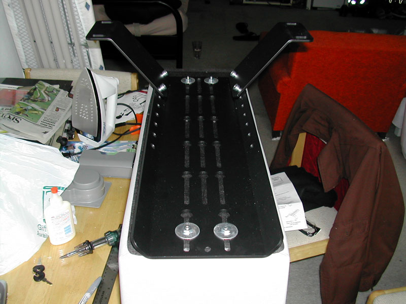 Black metal bracket with multiple bolt holes and two large bolts attached, resting on a white surface. Tools and papers are visible on the left