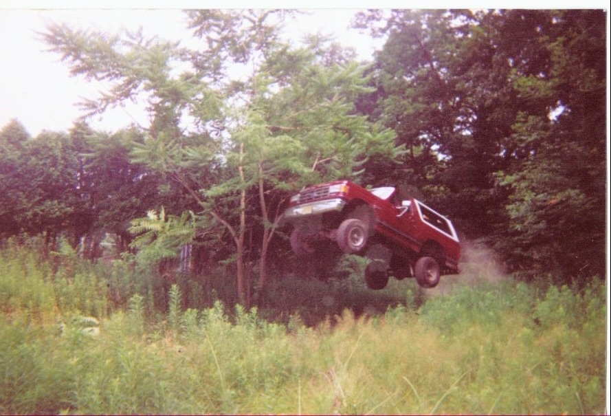 Red lifted Ford Bronco SUV jumping mid-air over grass in a wooded clearing