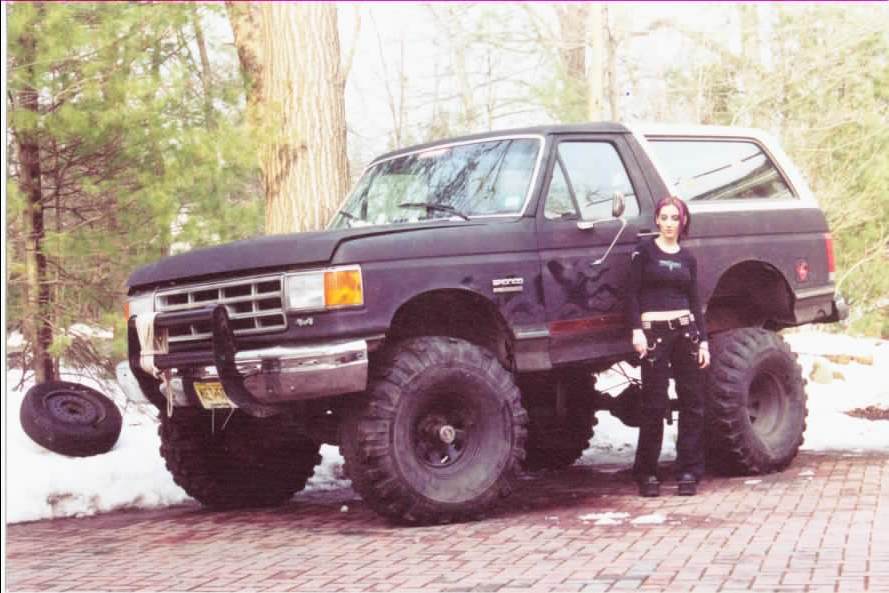 Lifted black Ford Bronco on big mud tires, person standing beside it on brick driveway with snow patches