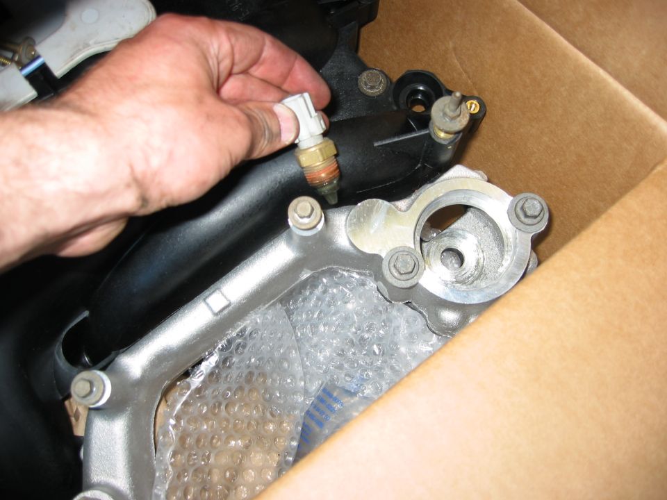 Hand holding a sensor on a metal engine component with black tubing and bubble wrap