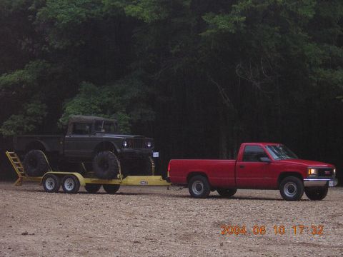 1984 Chevrolet Blazer - A red GMC pickup truck tows a dark off-road vehicle on a trailer through a gravelly area.