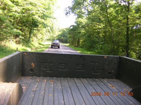1984 Chevrolet Blazer - View from the bed of a pickup truck shows a dark SUV ahead on a rural road surrounded by trees.
