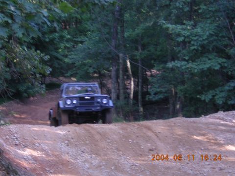 1984 Chevrolet Blazer - A dark off-road vehicle navigates a dirt path in a wooded area, with a timestamp and website logo visible in the lower right corner.