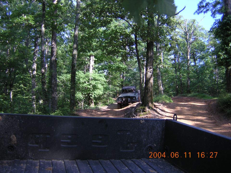 View from a truck bed looking at another off-road vehicle driving on a dirt path in a forest