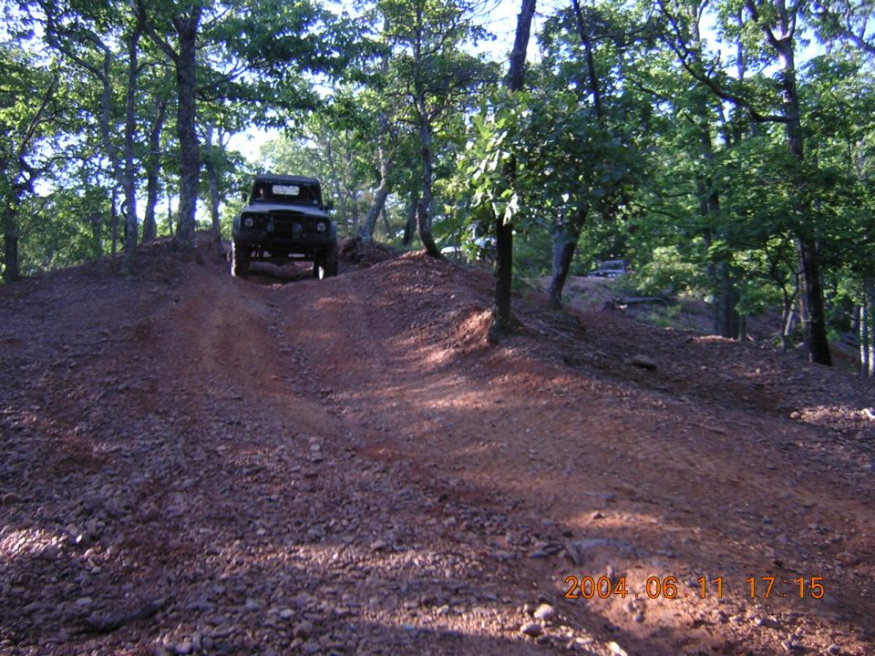 Jeep-like vehicle climbing a dirt hill in a wooded area. Another vehicle is visible in the background