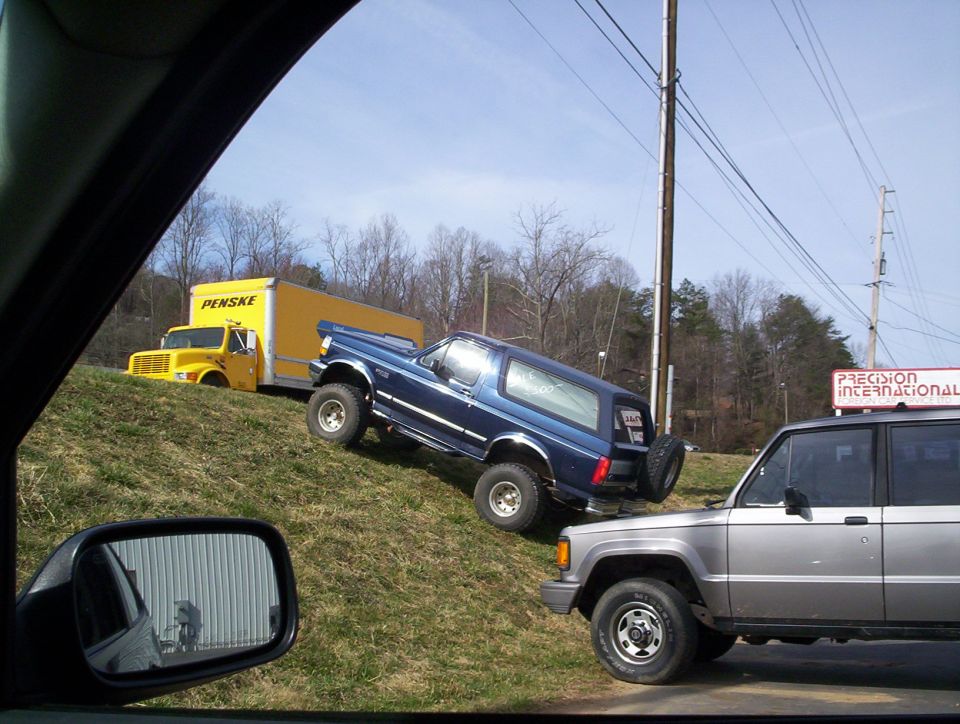 Blue lifted Ford Bronco parked on a grassy roadside slope beside a yellow Penske box truck