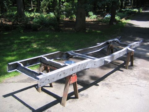 1978 Ford Bronco - A bare stripped truck frame sits upside-down on wooden sawhorses in a driveway, sandblasted or ground to clean bare metal with all crossmem…