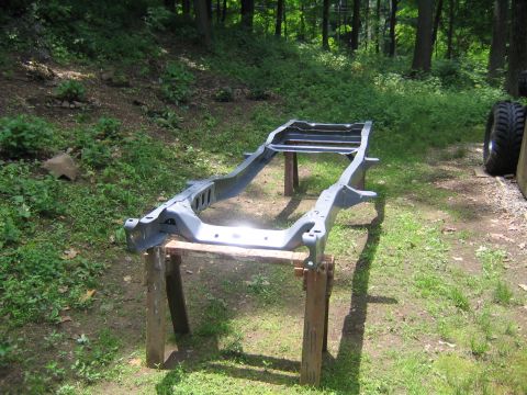 1978 Ford Bronco - Bare truck frame stripped of its body and freshly coated in gray, resting on wooden sawhorse stands in a grassy wooded yard during a frame-…