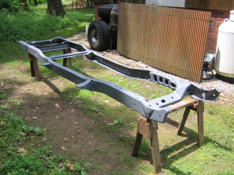 1978 Ford Bronco - A stripped truck ladder frame, freshly coated in dark gray primer or chassis paint, resting on wooden sawhorses in a grassy yard during a f…