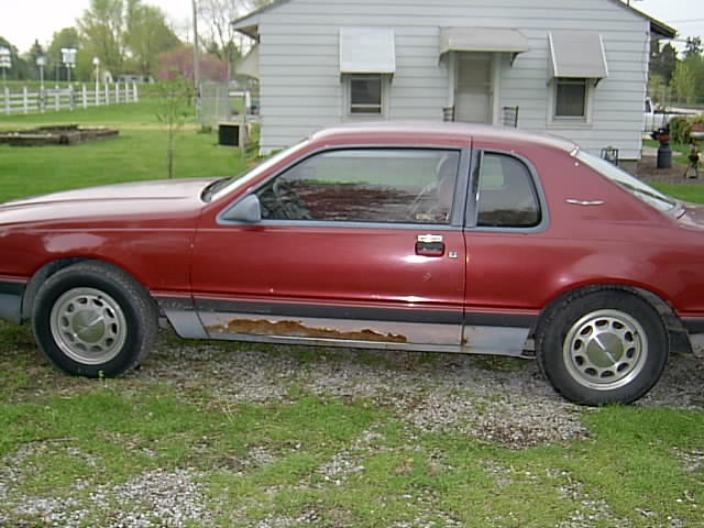 Side view of a rusty dark red two-door coupe parked on gravel by a white house