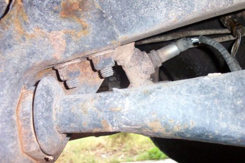 1995 Ford Bronco - Underside view of a rusty vehicle frame area showing an axle tube, U-bolt threads and mounting plate, with a brass fitting and a black flex…