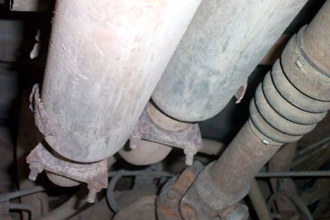 1995 Ford Bronco - Underbody view of a dual exhaust setup: two parallel gray mufflers secured with rusty U-bolt clamps, with the driveshaft, slip yoke, and U-…