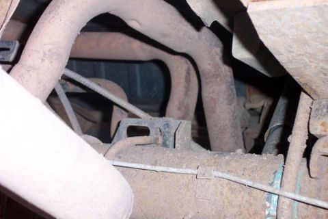 1995 Ford Bronco - Underbody view of dust-coated exhaust piping curving over a crossmember beneath a vehicle, with surface rust on the pipes and nearby bracke…