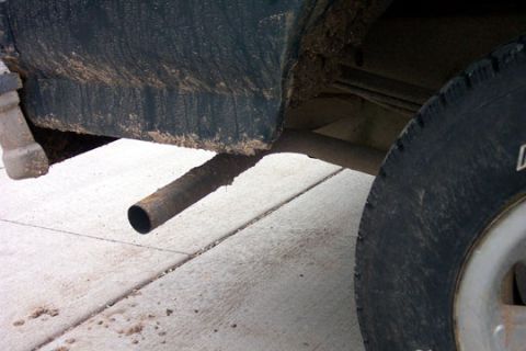 1995 Ford Bronco - Close-up under the rocker panel of a mud-caked dark truck showing a plain steel exhaust tailpipe angled down toward the concrete driveway j…