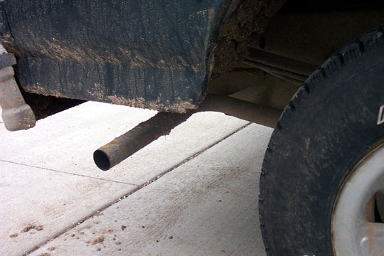Downturned steel exhaust tailpipe under a muddy truck rocker panel beside an all-terrain tire