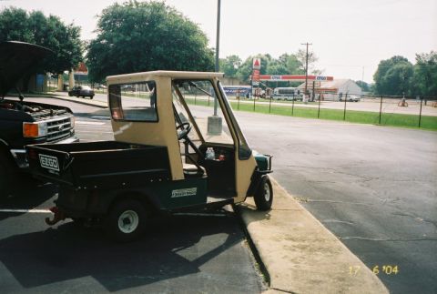 1988 Ford Bronco - A utility golf cart is parked next to a truck with its hood open, with a gas station in the background.
