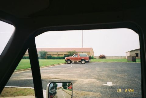 1988 Ford Bronco - Side view of a red 1988 Ford Bronco parked in an asphalt lot with buildings in the background.
