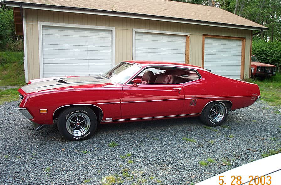 Red 1970 Ford Torino fastback with hood scoop and chrome wheels parked by a garage