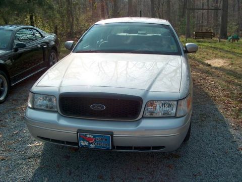 2000 Ford Crown Victoria - A silver Ford Crown Victoria is parked in a gravel driveway next to a black car, with trees in the background.