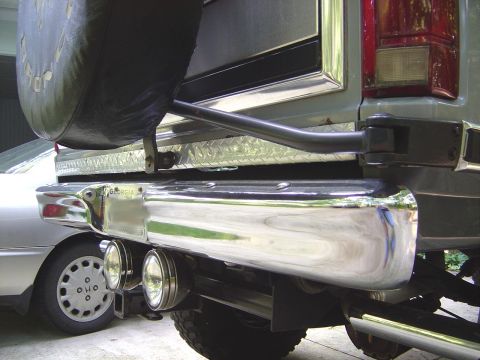 1986 Ford Bronco - Close-up of the rear corner of a silver Ford Bronco in a garage, showing a polished chrome rear bumper with two round driving lights, a dia…