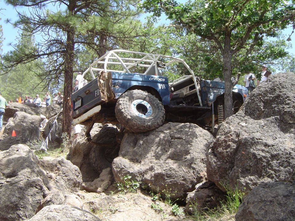Blue tube-cage buggy climbing a stack of large boulders at a forested rock-crawling event