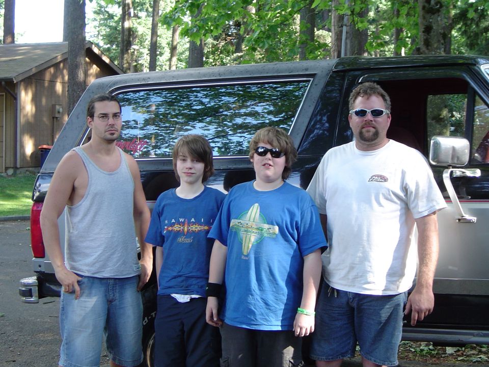 Four people standing in front of a dark SUV with chrome bumper in a wooded campsite