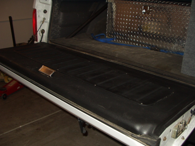 Black tailgate laid open with chrome trim, diamond-plate toolbox behind in garage.