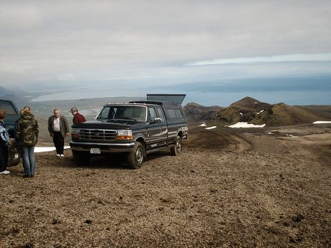 1996 Ford Bronco - A group of people stand near a pickup truck in a , scenic landscape with mountains and a body of water in the distance.