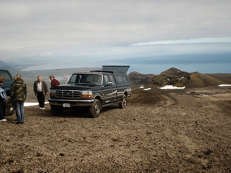 Dark Ford pickup with camper shell parked on gravel with people and coastal mountains behind