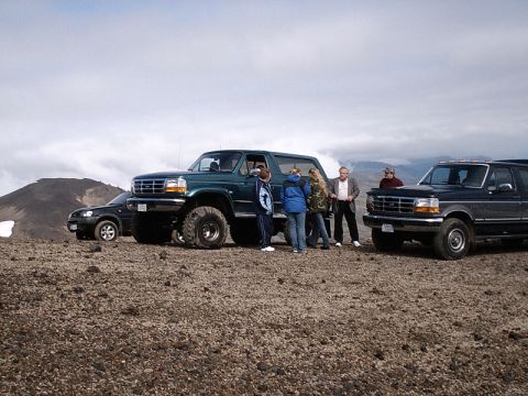 1996 Ford Bronco - A group of people stand around a lifted green 1996 Ford Bronco and a dark SUV on a rocky, barren landscape.