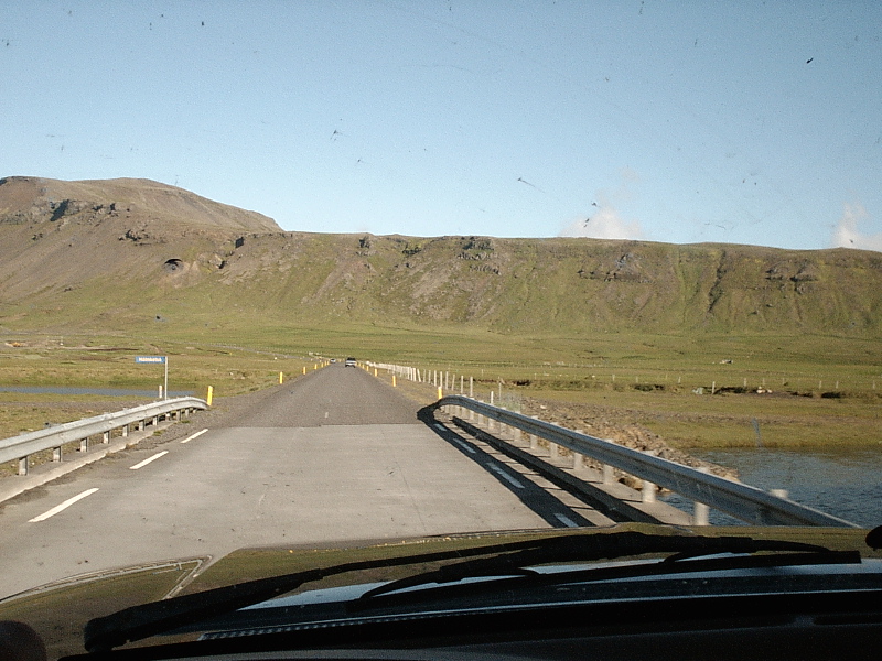 Driver's-eye view crossing a narrow bridge toward green hills under clear sky.