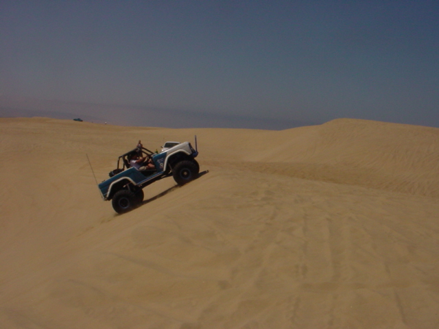 Blue and white open-top Jeep CJ climbing a tan sand dune under hazy sky
