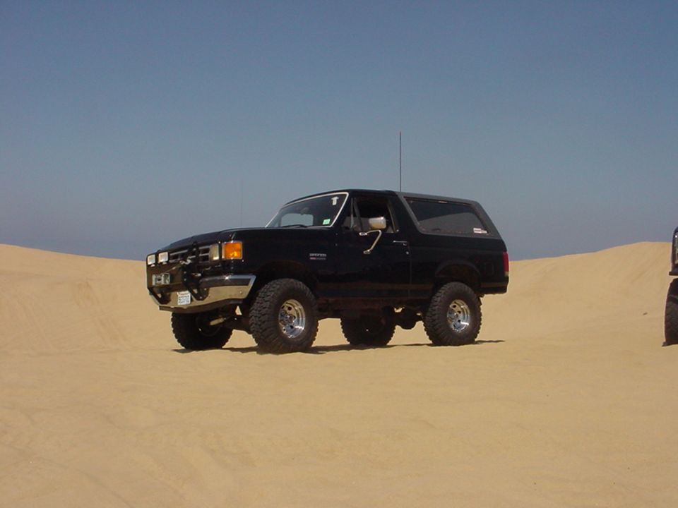 Black full-size Ford Bronco with chrome wheels parked on a sand dune under blue sky