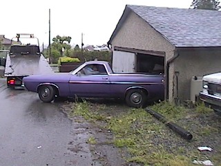 1968 Ford Ranchero - A purple pickup truck, likely from the 1970s, is parked beside a garage, with a tow truck visible in the background.