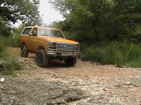 1986 Ford Bronco - A yellow SUV navigates a rocky, off-road trail surrounded by trees and tall grass.