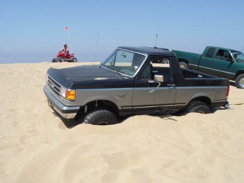 1989 Ford Bronco - A two-tone 1989 Ford Bronco is stuck in the sand at the beach, with an ATV and another pickup truck visible.