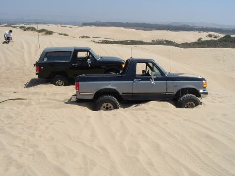 1989 Ford Bronco - Two 1989 Ford Broncos are stuck in sand dunes on a sunny day.