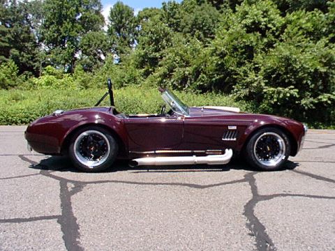 1967 Ford 427 - This dark red convertible sports car has side pipes and black wheels, with trees in the background.