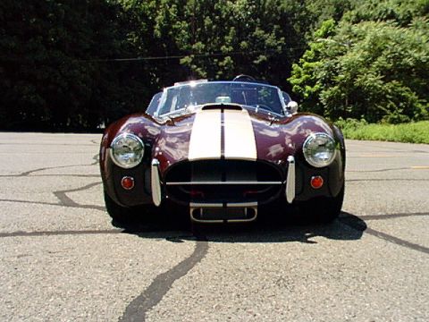 1967 Ford 427 - A classic dark red convertible with cream racing stripes is parked on an asphalt road, facing the camera.